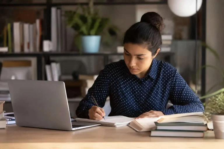Focused student writing notes at a desk with laptop and books in a quiet indoor study setup